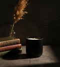 Stone Manor candle in matte black vessel on weathered stone with leather books and pampas grass